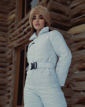 Woman in warm white clothing posing confidently outdoors during winter, with wooden building backdrop.