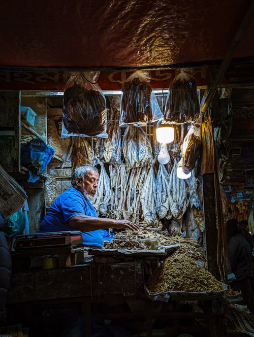 Free An elderly man in a blue shirt at a market stall selling dried fish. Warm lighting creates a cozy atmosphere. Stock Photo