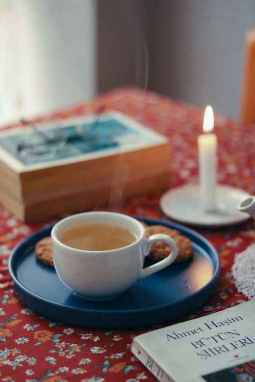 Free Warm coffee cup on tray with cookies, beside candle and books on a patterned tablecloth. Stock Photo