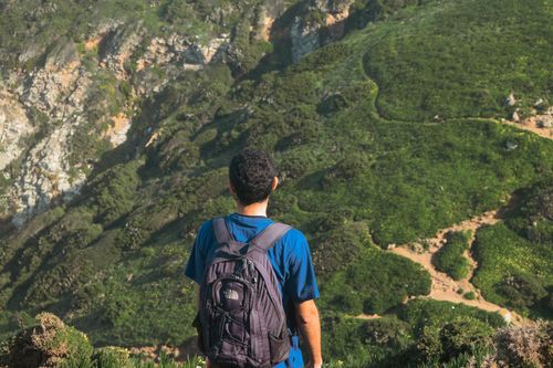 Free Man with backpack stands facing lush green mountain scenery, embracing nature's beauty. Stock Photo