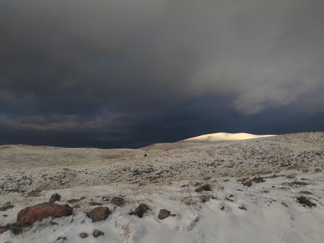 A snowy winter landscape with hilly terrain under a dramatic dark sky, showcasing nature's raw beauty.