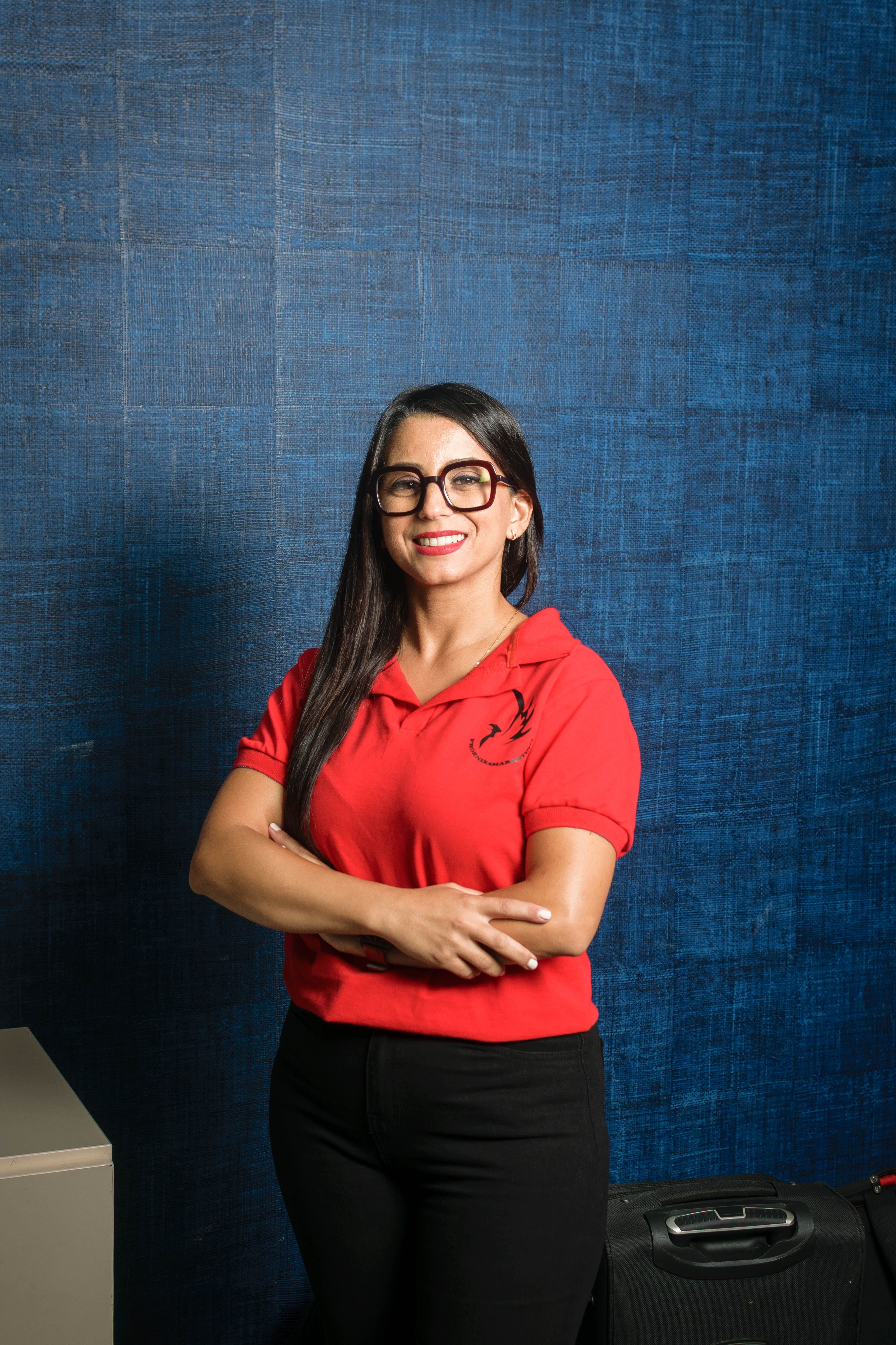 A young woman in a red top poses confidently against a blue background in a studio setting.