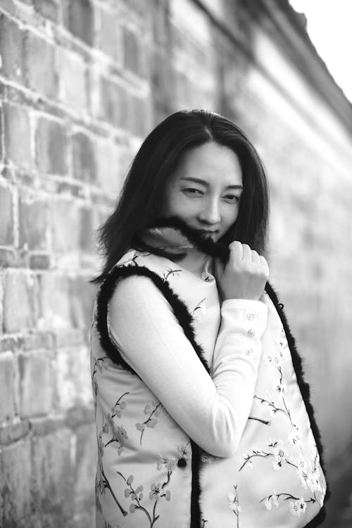 A woman in a vest and jacket leaning against a brick wall