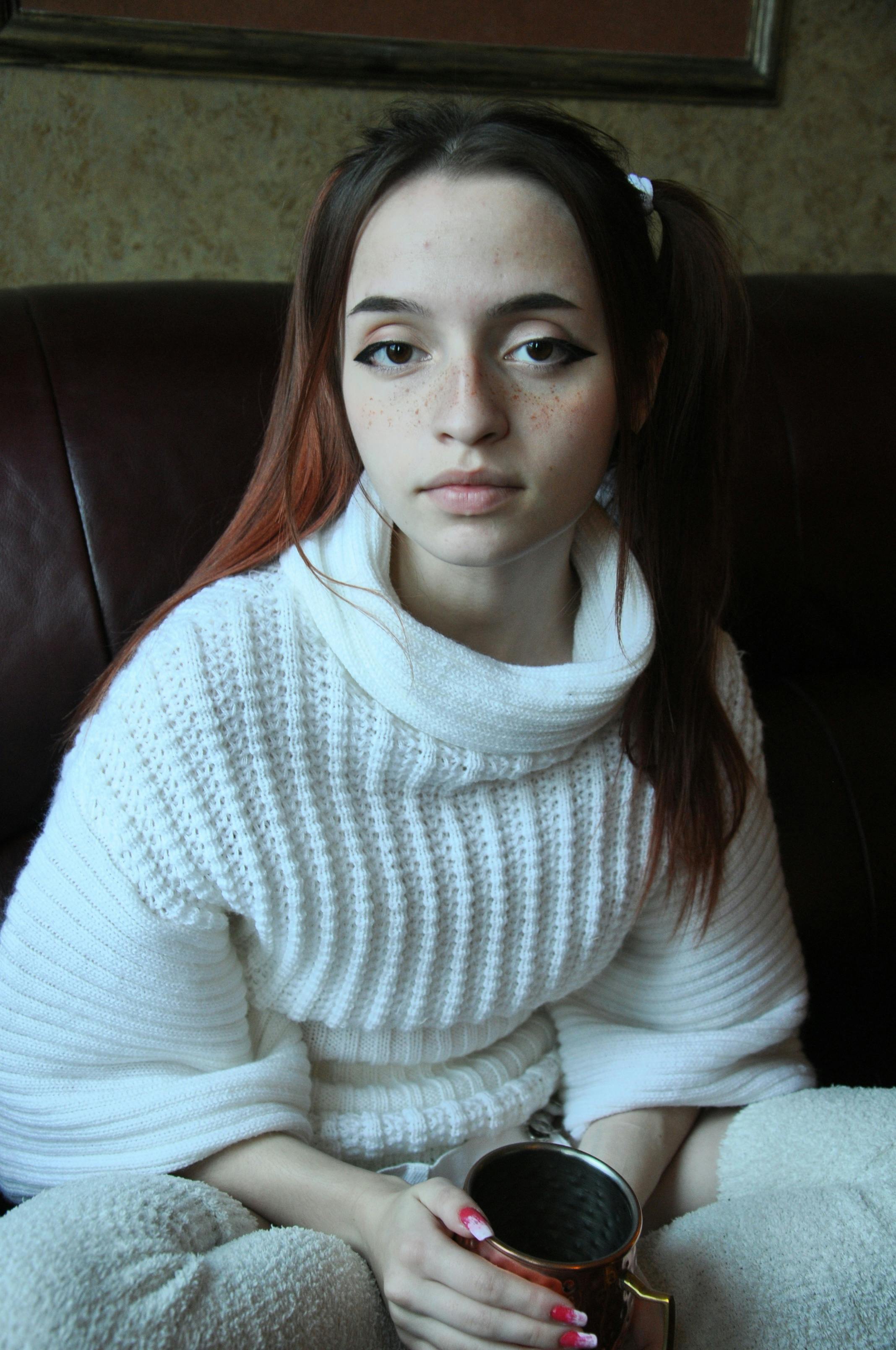 Portrait of a young woman in a white sweater holding a cup indoors, conveying a cozy atmosphere.