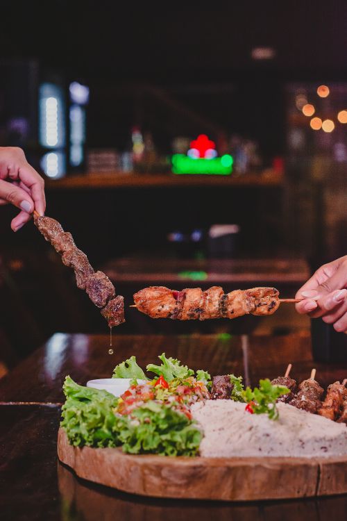 Free Close-up of hands serving meat skewers on a wooden board with lettuce garnish. Stock Photo