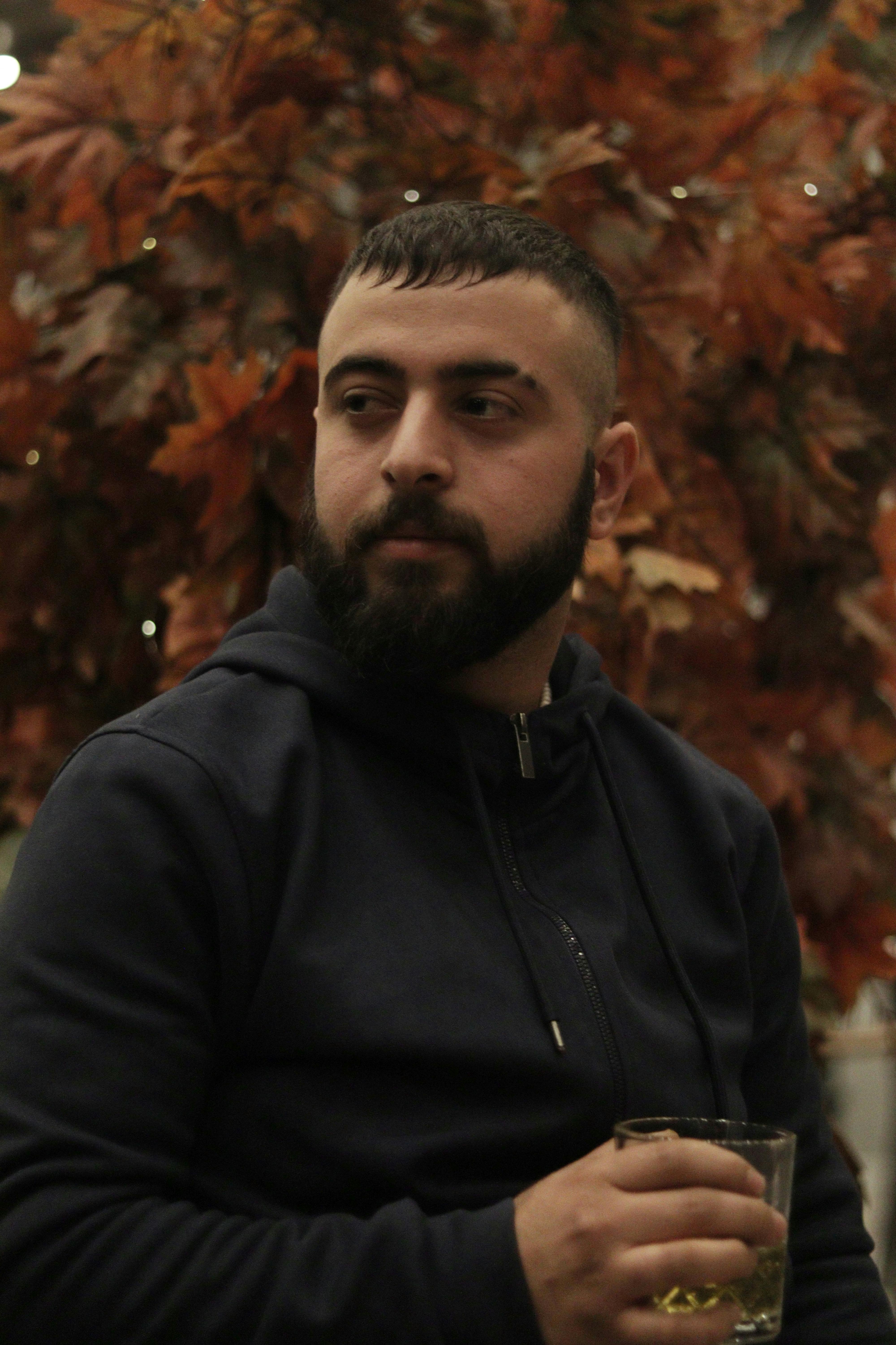 A serious, bearded man in a black hoodie sits with a drink in hand during fall.