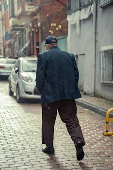 Rear view of an elderly man in a blue jacket walking down a city street on a rainy day.