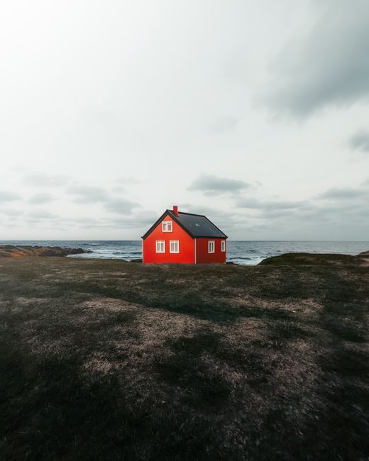 A striking red house on a grassy shore against a cloudy sky and ocean backdrop.