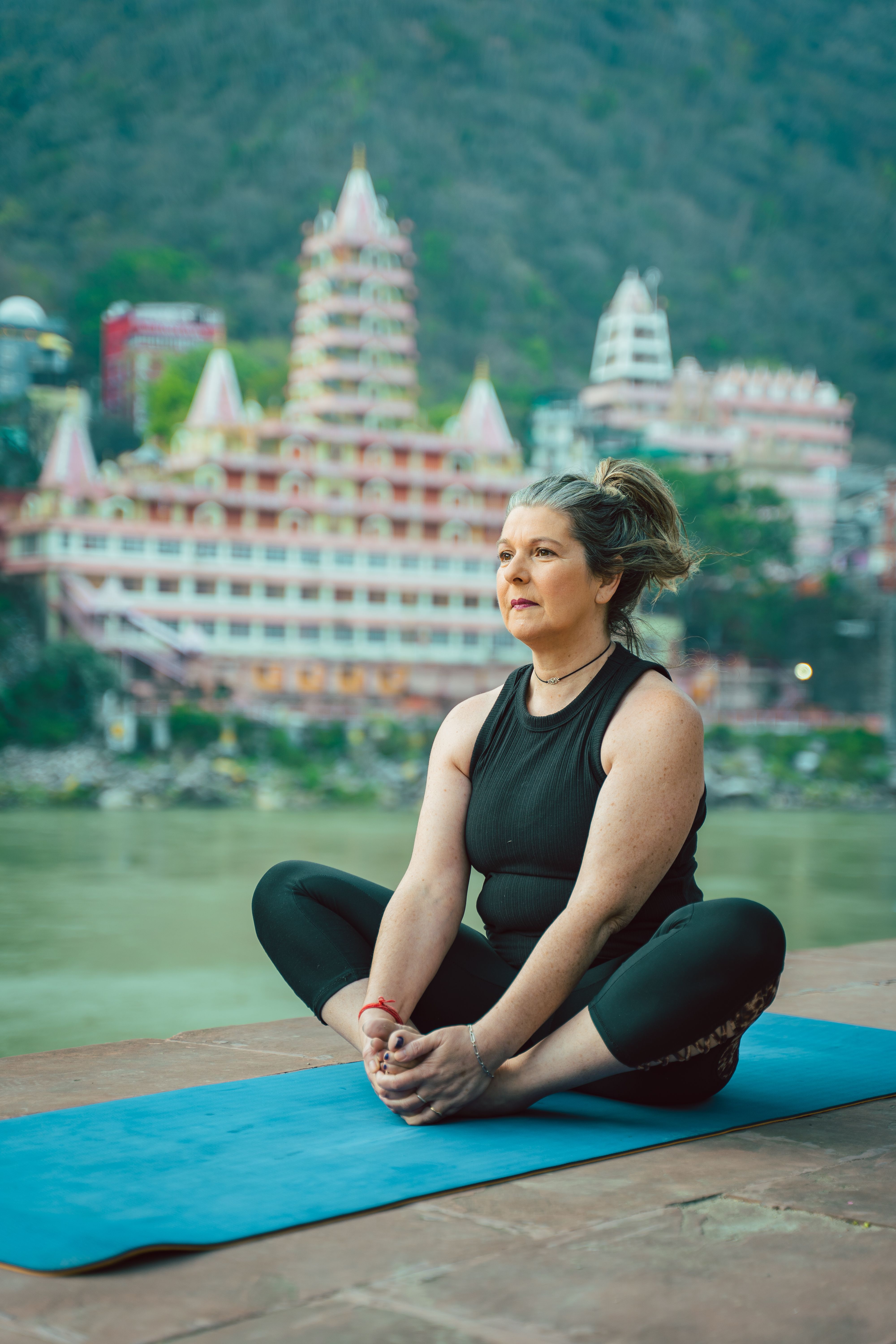 Adult woman doing yoga on a mat by the river with iconic Rishikesh skyline in the background.