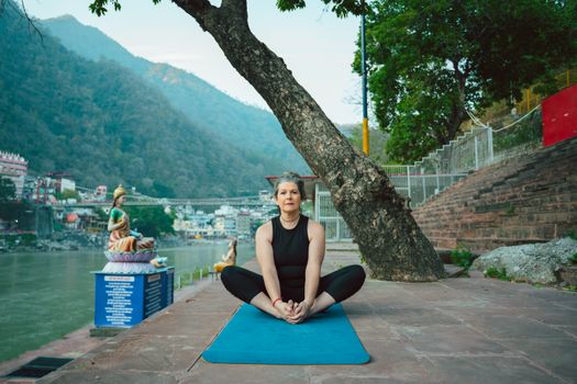 Woman practicing yoga outdoors by the Ganges River in Rishikesh, India with mountains in the background.