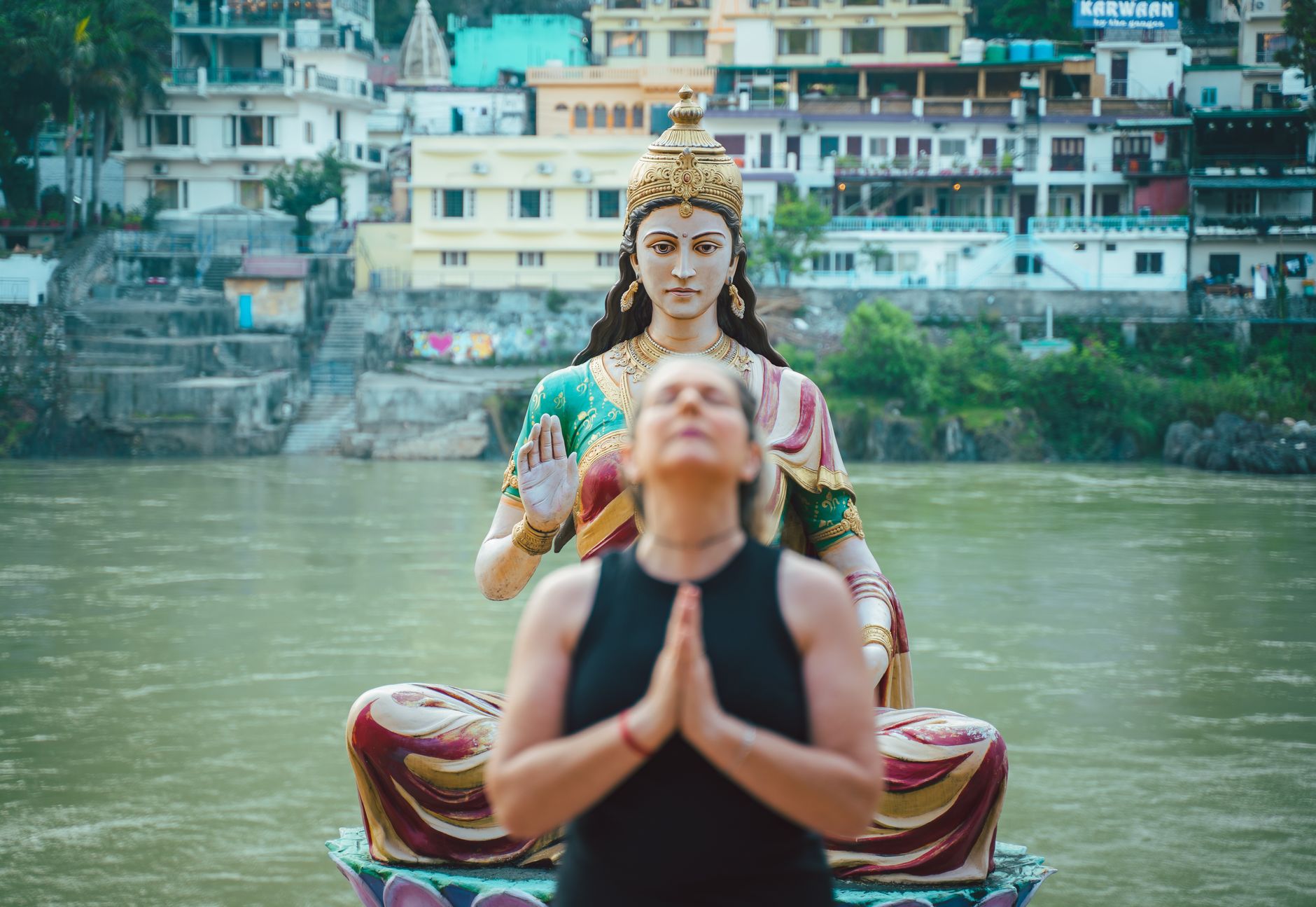 Woman practicing yoga in front of a statue by the riverside, surrounded by buildings.