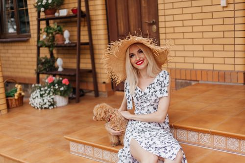 Free Smiling woman in floral dress and straw hat with small dog sitting outdoors. Stock Photo