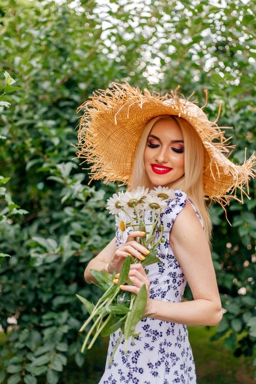 Free Blonde woman in a sundress holding daisies, smiling, outdoors in a lush garden. Stock Photo
