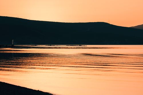 Free A tranquil sunset casts a golden glow over a calm lake with gentle ripples and a silhouetted hill. Stock Photo