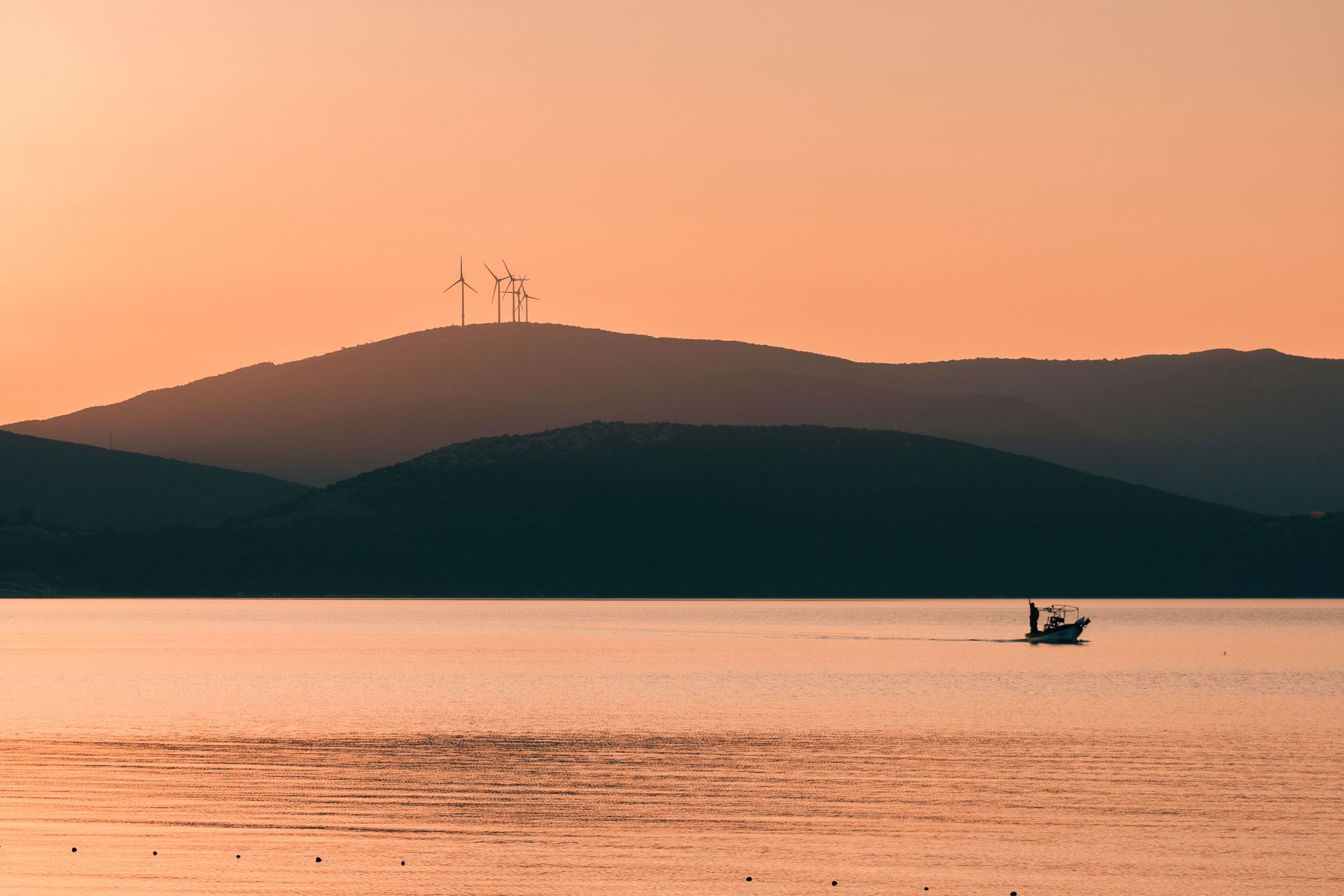 Silhouette of a boat on a calm sea at sunset with wind turbines atop distant hills.