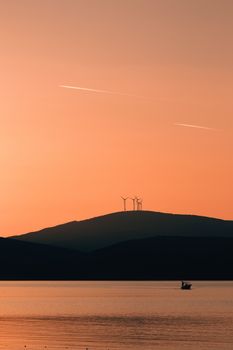 Beautiful sunset silhouette of hills with wind turbines and a calm lake view.