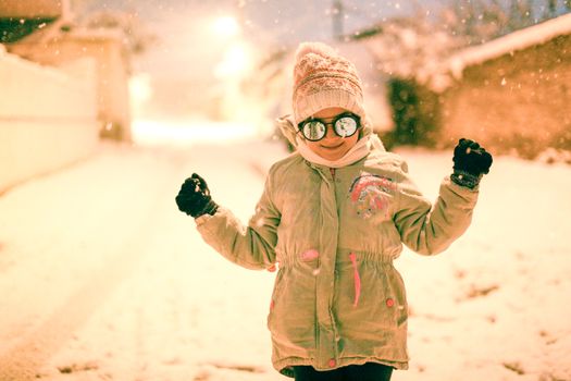 A joyful child wearing a winter coat and hat, enjoying snowfall in an urban street.