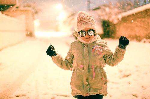 Free A joyful child wearing a winter coat and hat, enjoying snowfall in an urban street. Stock Photo