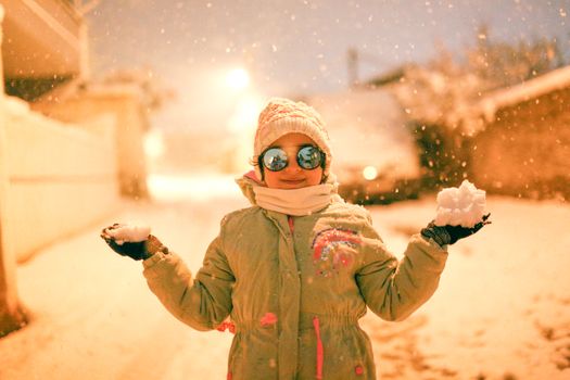 A cheerful child in winter attire enjoying the snowfall in a city street at night.