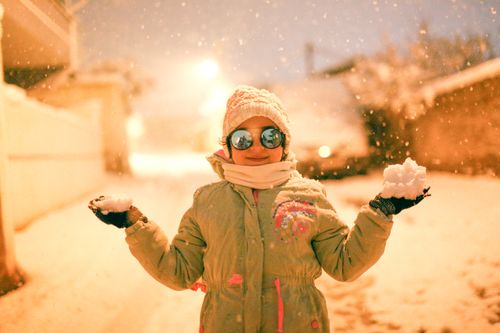 Free A cheerful child in winter attire enjoying the snowfall in a city street at night. Stock Photo