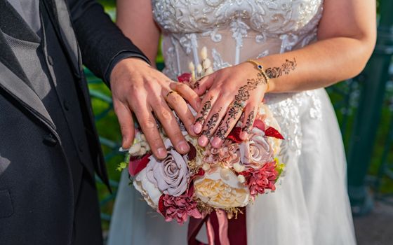 Close-up of a bride and groom's hands with rings and henna, elegantly holding a floral bouquet.