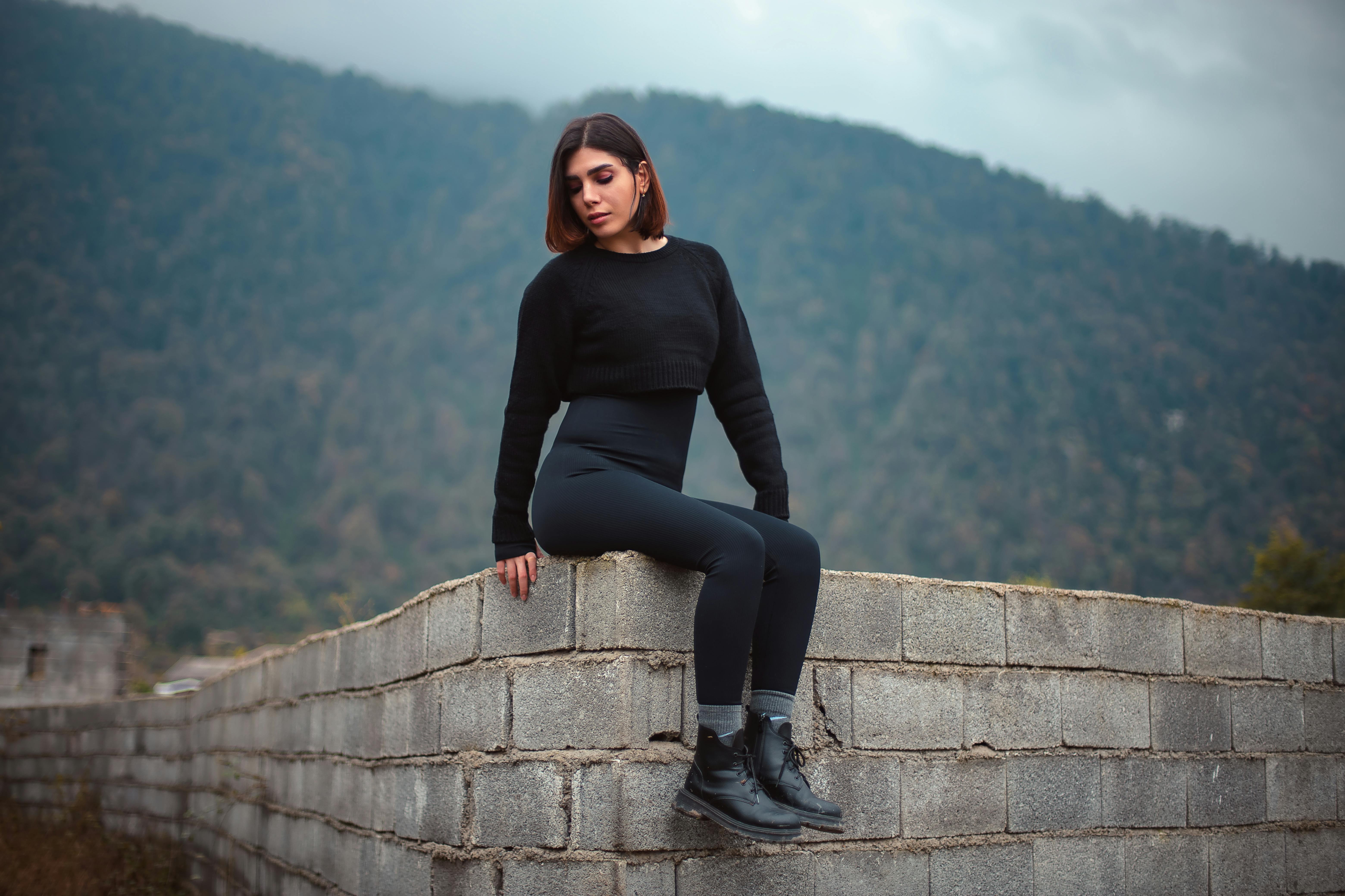A stylish woman in black clothing poses on a stone wall against a scenic mountain backdrop.