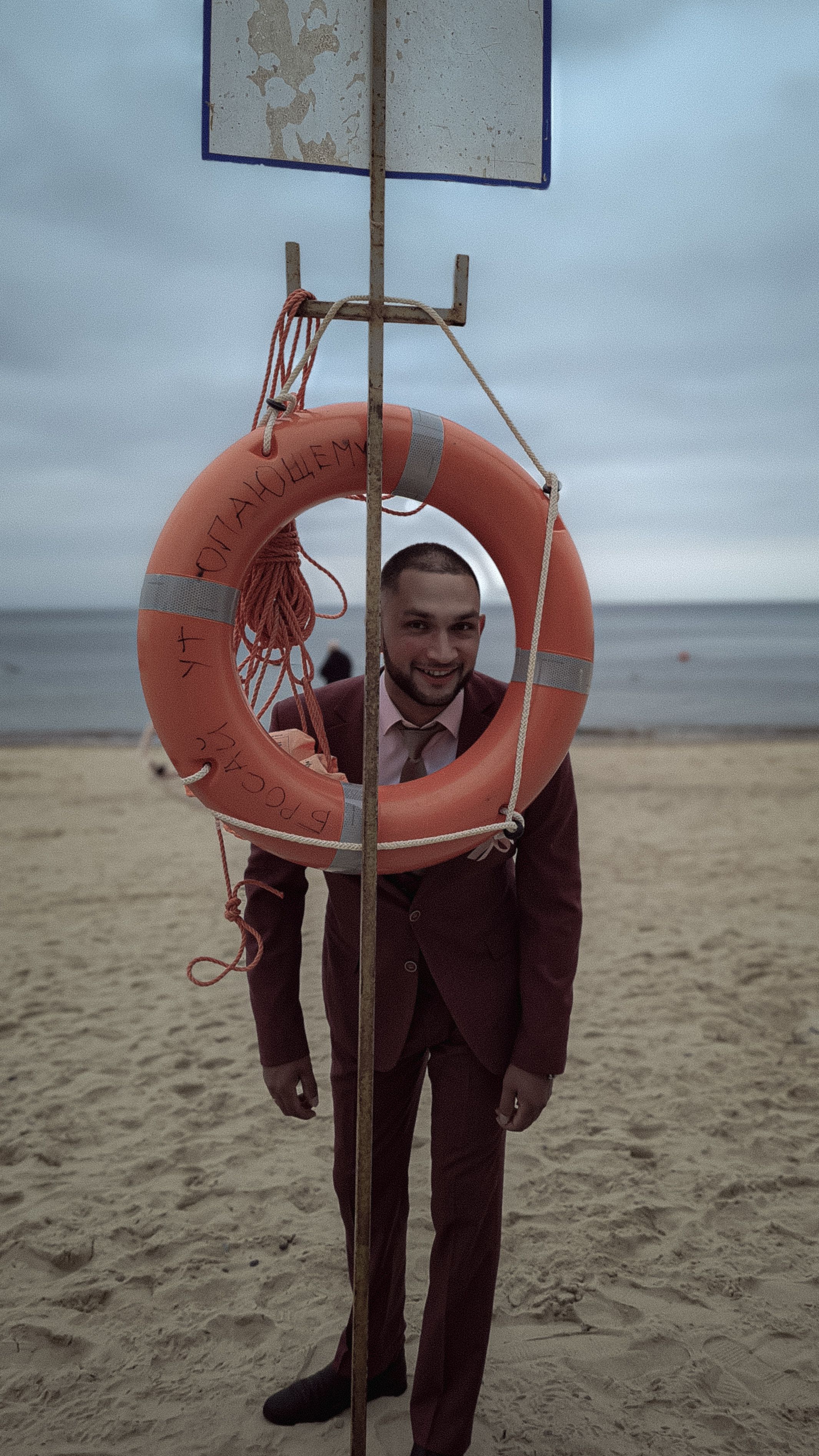 A cheerful man in a suit stands by a lifebuoy on a sandy beach with an overcast sky.