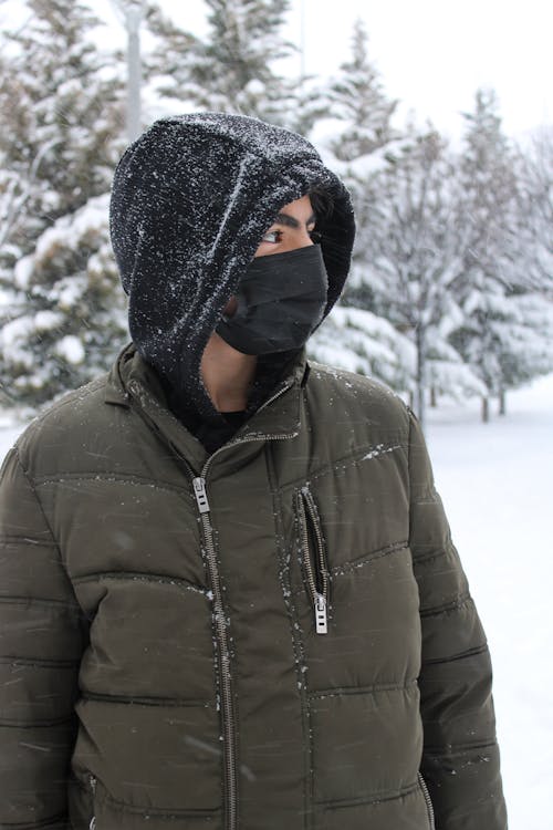 Man in a Hoodie and Jacket Wearing a Face Mask while Standing on a Snowy Field 