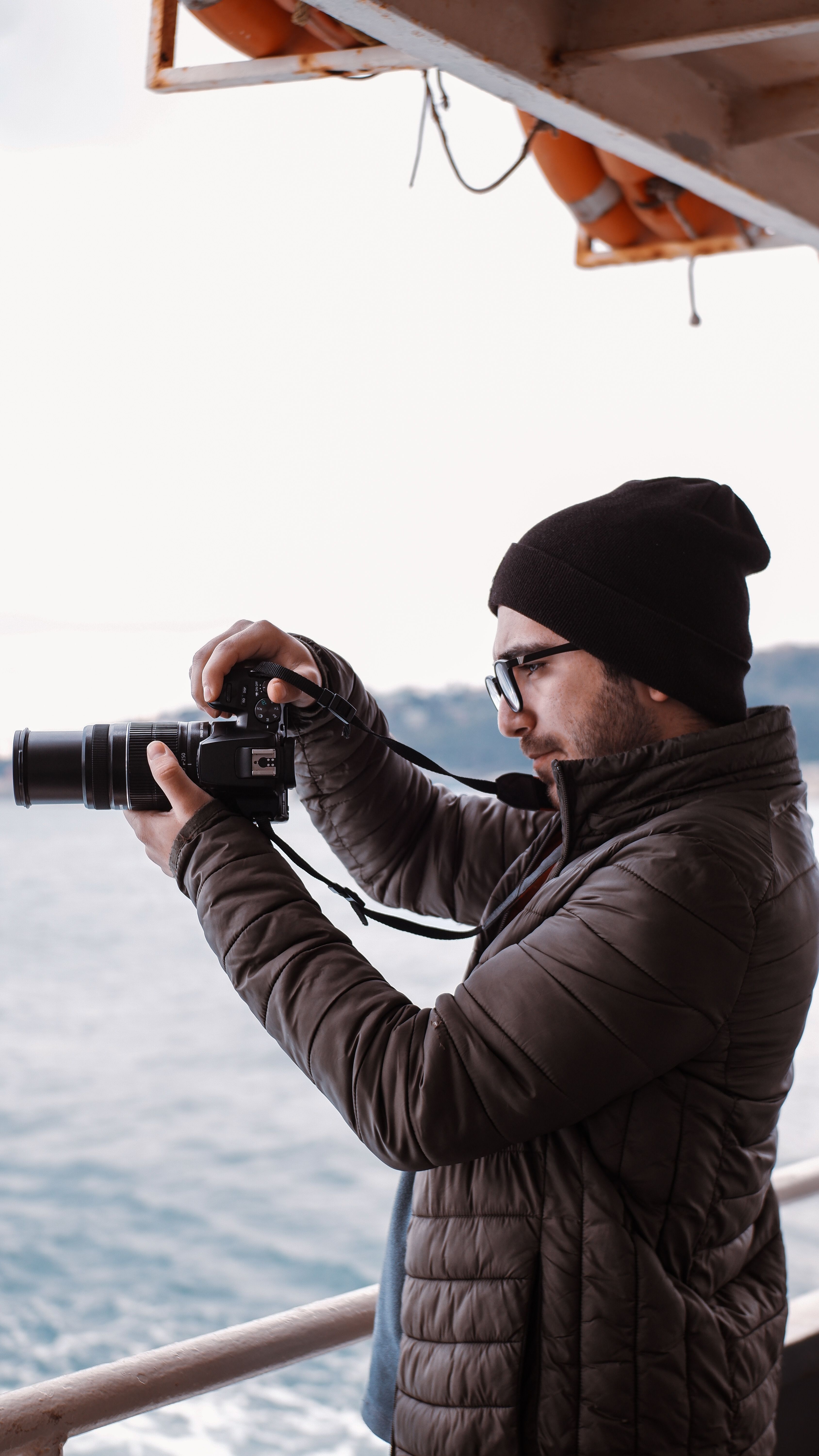 A man in a black jacket and beanie taking photos by the sea on a cool day.