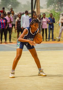 Focused teenage girl playing basketball in sportswear with onlookers.