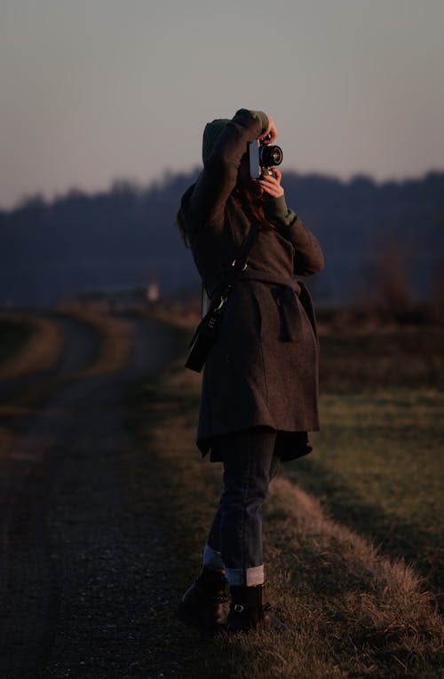 Woman in Black Coat Taking Pictures on Dirt Road