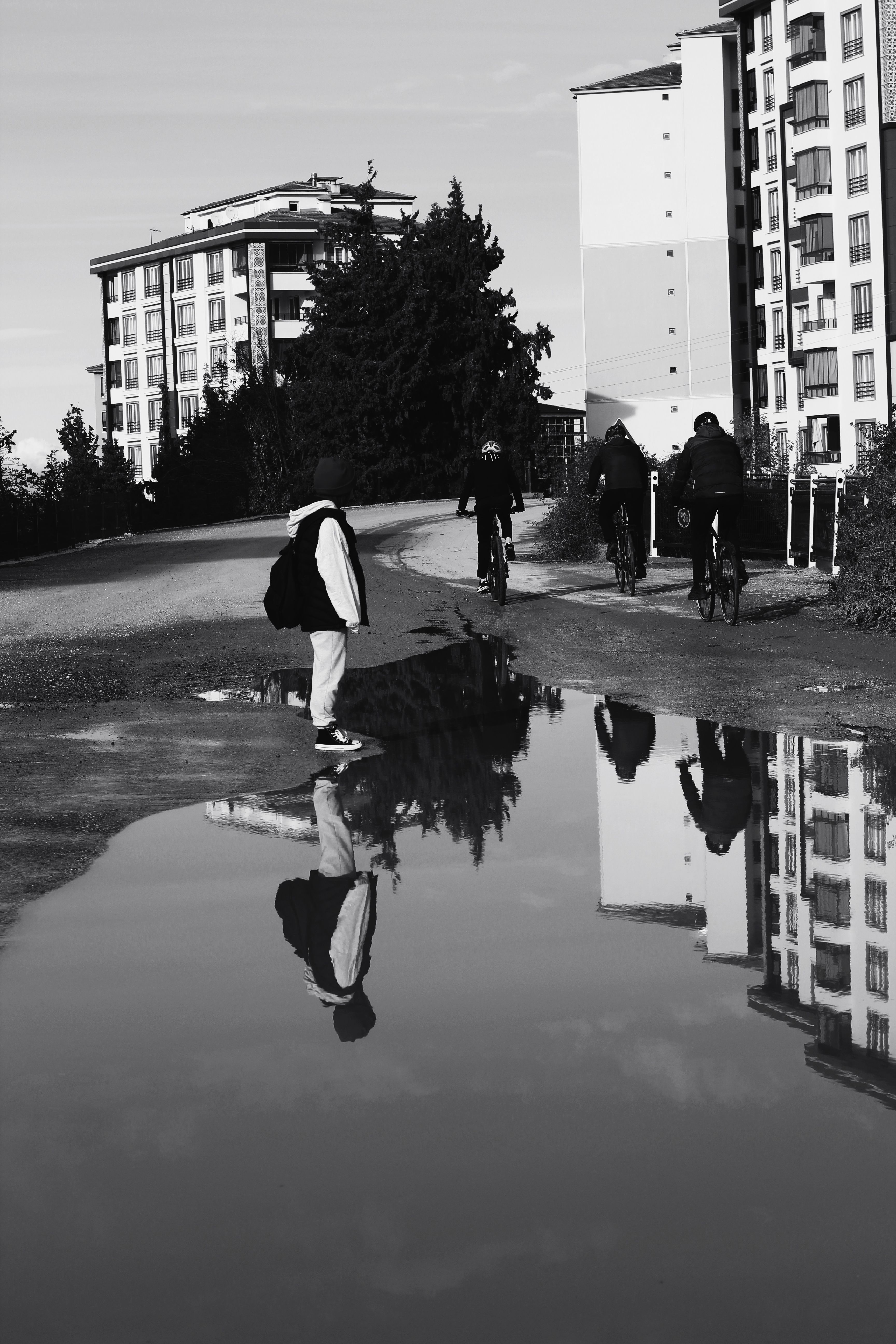A monochrome cityscape featuring people and cyclists reflecting in a puddle.