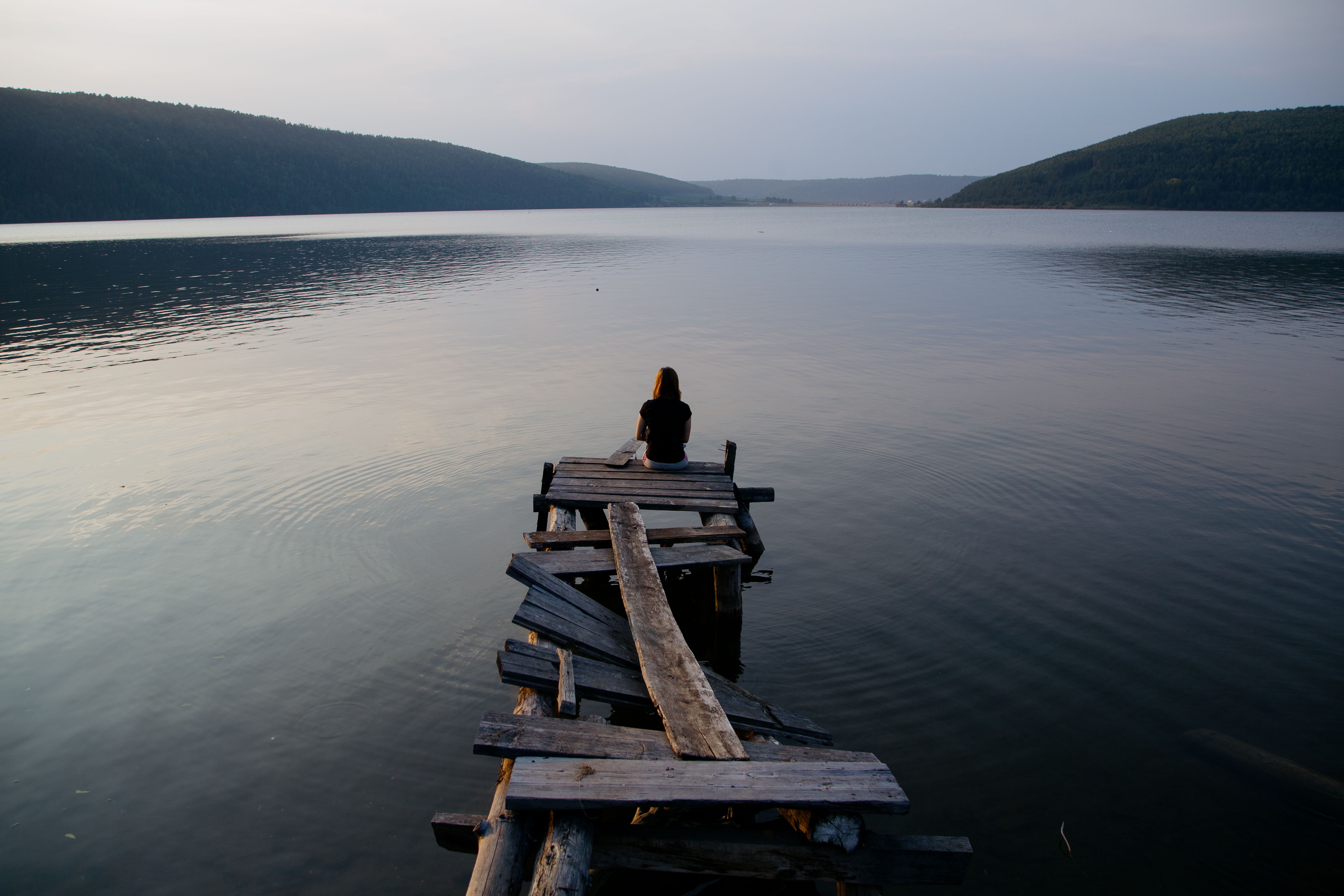 A solitary woman sits on a weathered pier, gazing across a serene lake at sunset, inviting contemplation.