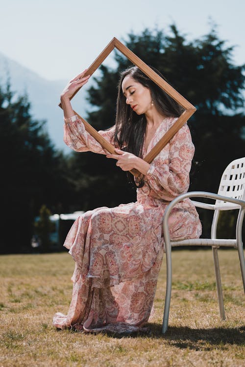 Woman Posing in Floral Maxi Dress Sits on Chair with Painting Frame