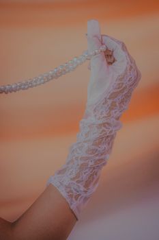 Close-up of a bridal hand in lace gloves holding a pearl necklace against a warm orange backdrop.