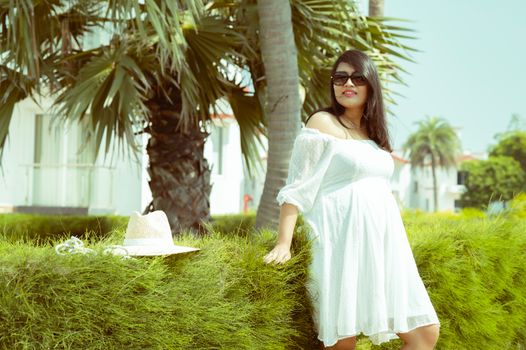 Pregnant woman in white dress enjoying a sunny day outdoors by a hedge.