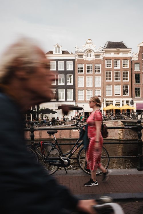 Free Blurred cyclist passing by iconic Amsterdam canal houses with a pedestrian in focus. Stock Photo