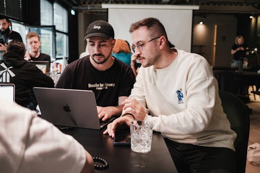 Two men collaborating on a project in a modern café setting with laptops and glasses.