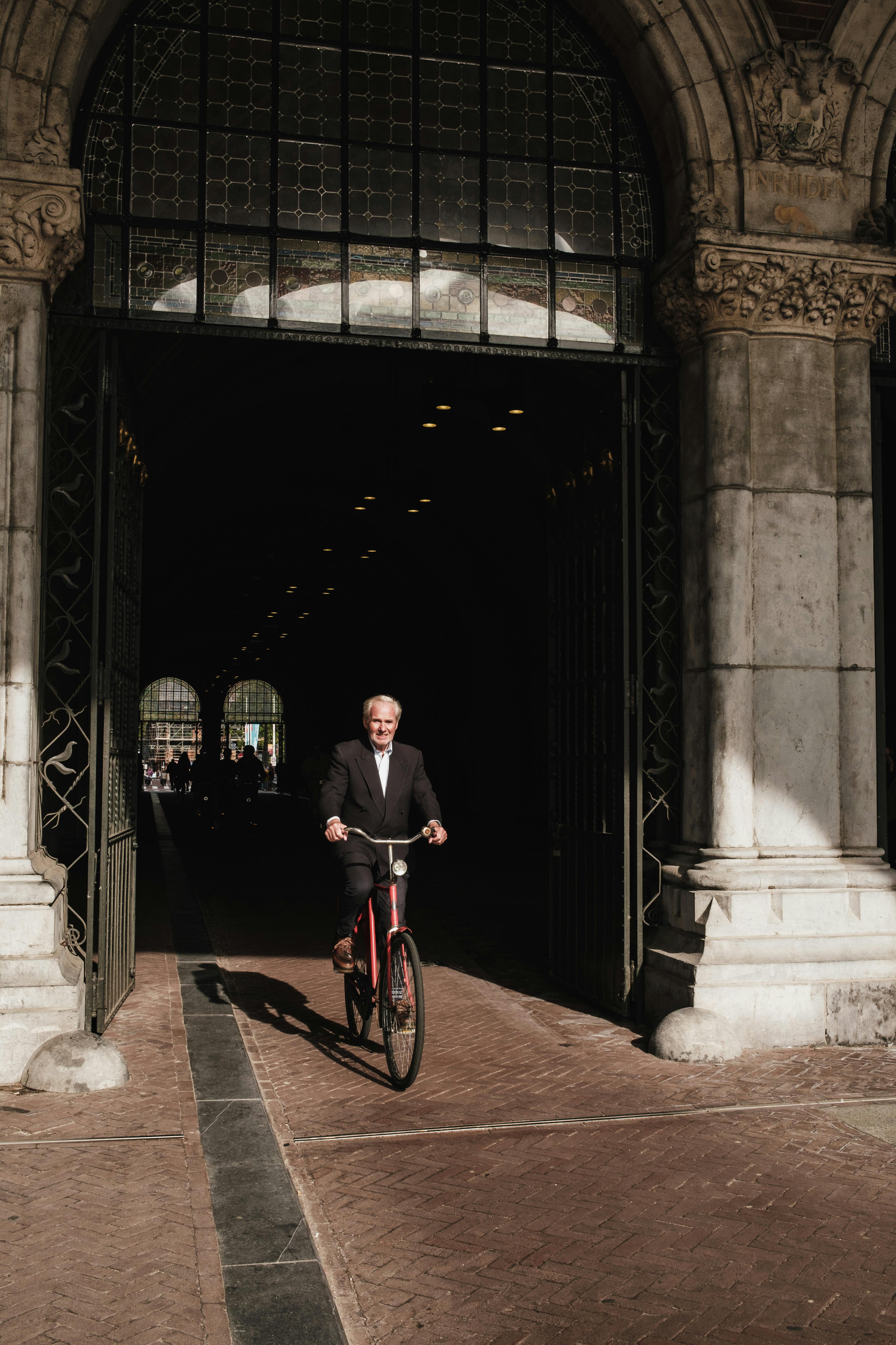 An elderly man in a suit cycling through a historic archway in an urban setting.