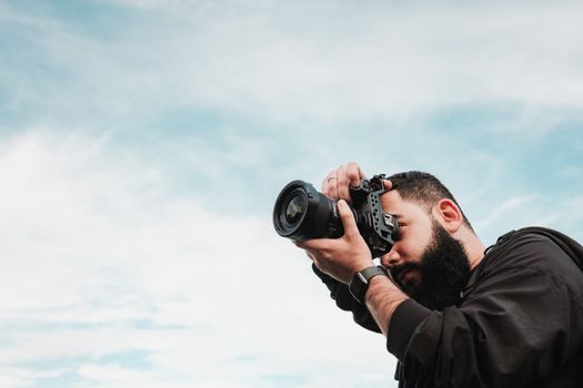 Portrait of a bearded photographer capturing outdoor scenes with a digital camera under a cloudy sky.