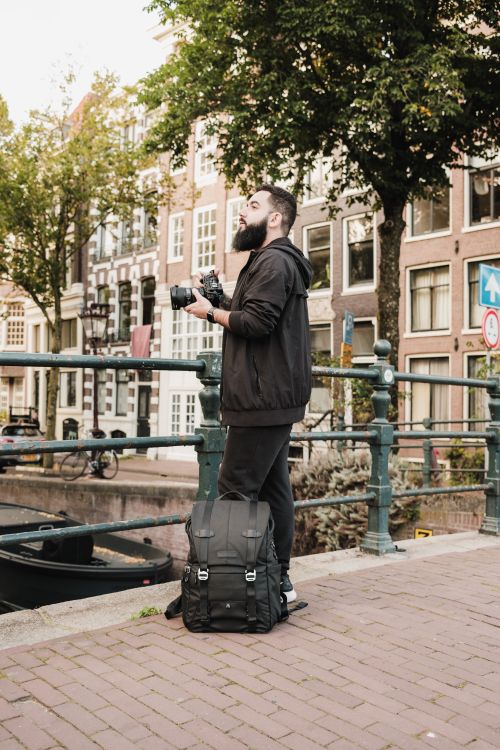 Free Bearded man with camera stands by Amsterdam canal, appreciating the urban scenery. Stock Photo