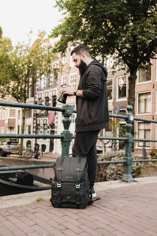 Free Young man with backpack and camera photographing a canal scene in Amsterdam. Urban exploration and travel photography. Stock Photo