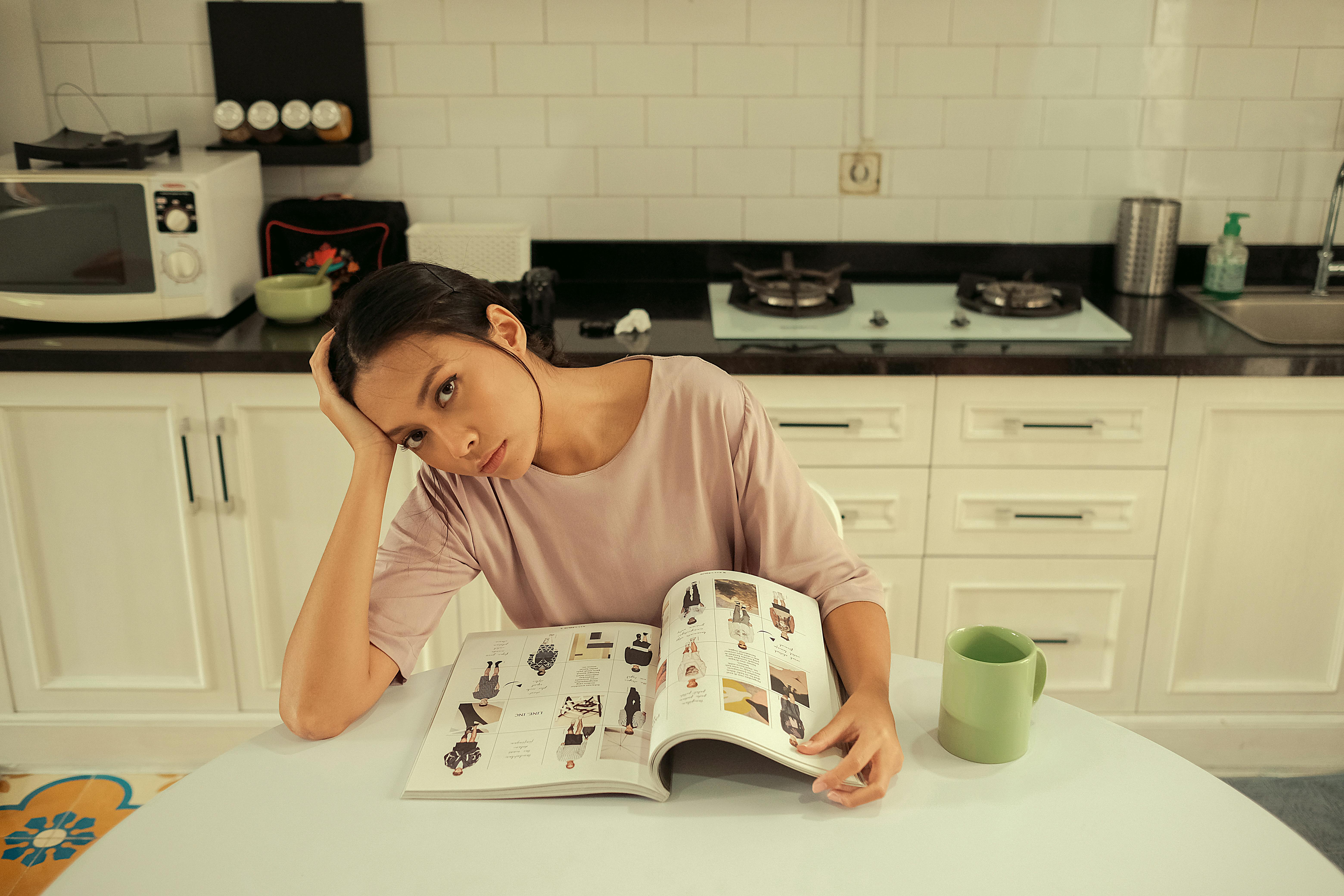 Woman sitting in a kitchen, reading a magazine at a table with a thoughtful expression.