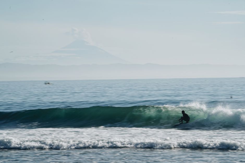 A surfer skillfully rides ocean waves with a stunning volcano backdrop, captured during daylight.
