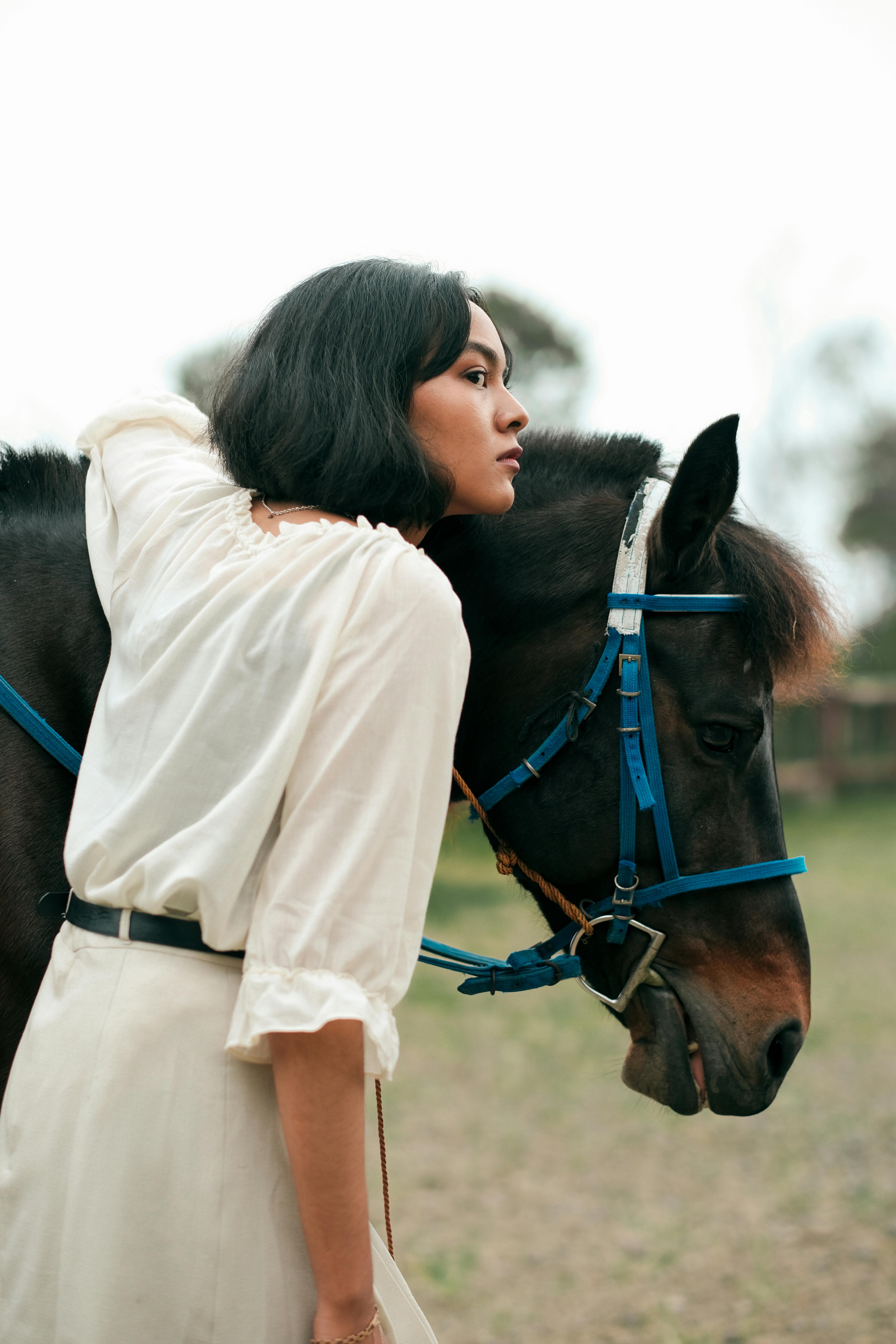 A woman in a white blouse embraces a horse with a blue bridle on a rural farm.