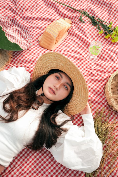 Young Woman in a Straw Hat and White Blouse Lying on a Plaid Picnic Blanket