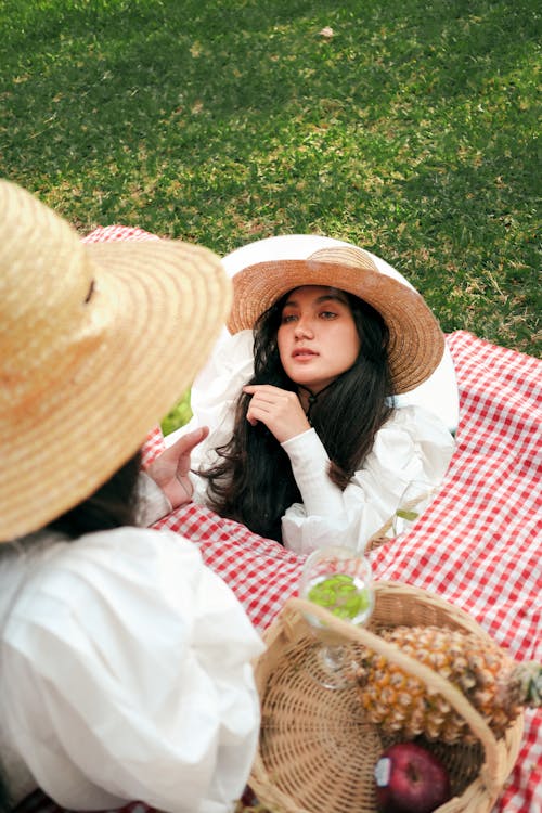 Young Woman in a Straw Hat Looking in the Mirror Sitting on a Picnic Blanket