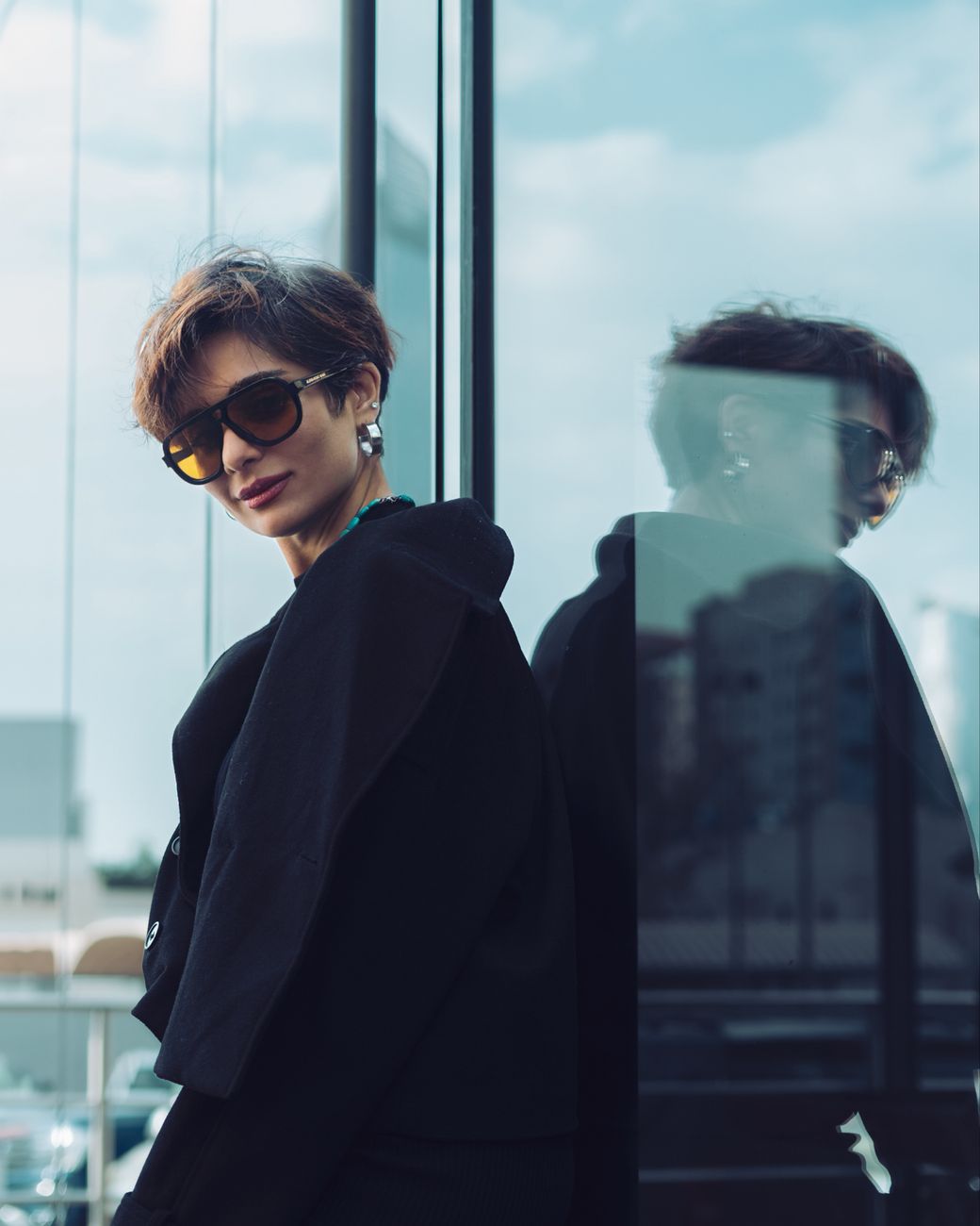 Woman in black coat and sunglasses posing in a modern cityscape with a glass reflection.