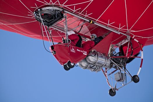 A vibrant red hot air balloon soaring through a clear sky with two passengers onboard.