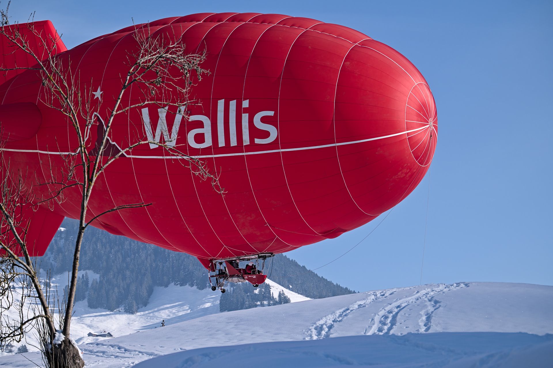 A vibrant red airship named Wallis flying over snow-covered hills in Switzerland under a clear blue sky.